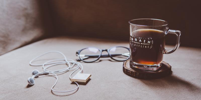 Kuvituskuva: Kahvikuppi jossa kahvia, langalliset kuulokkeet ja silmälasit.