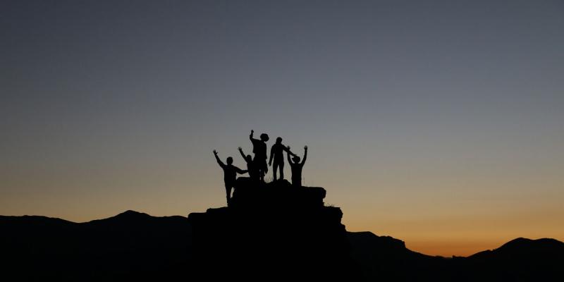 Viisi henkilöä kädet ilmassa kalliolla piirtyy aamutaivasta vasten.